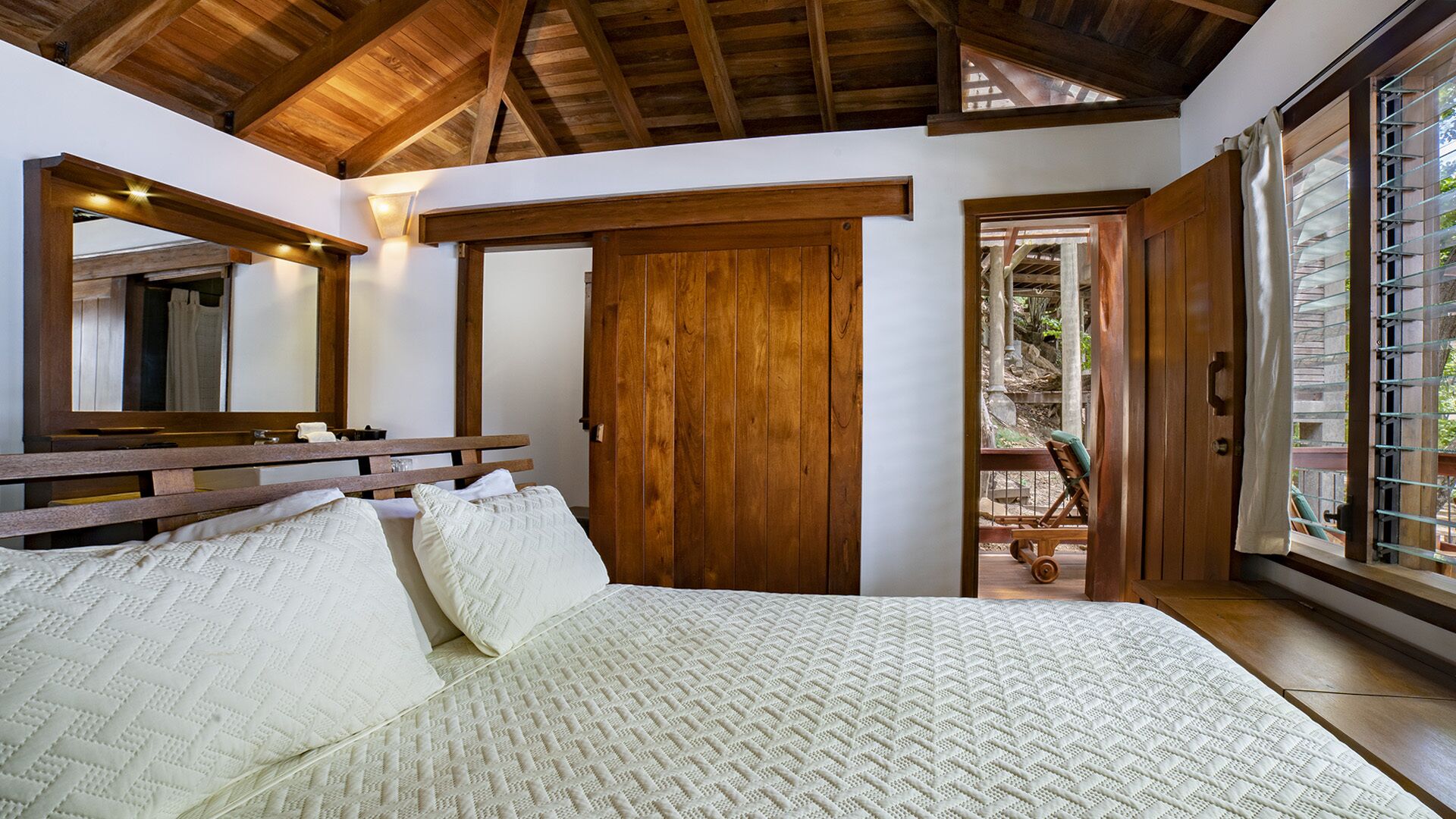 Camera da letto con soffitto in legno, un letto grande con biancheria bianca, un armadio in legno e una porta scorrevole in vetro che dà su una zona esterna.