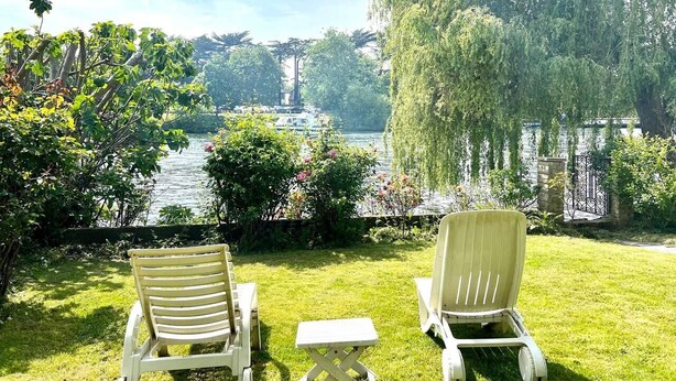 Two outdoor chairs facing a lake with a weeping willow tree and a stone wall in the background.