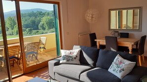 Ein Wohnzimmer mit einer Couch, einem Esstisch mit Stühlen, einem großen Spiegel und Blick auf einen Balkon mit Holzstühlen.