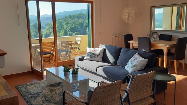 Ein Wohnzimmer mit einer Couch, Sesseln, einem Couchtisch und Blick auf einen Balkon mit Tisch und Stühlen.