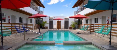 Une piscine entourée d’une clôture en bois et d’un bâtiment blanc doté de balcons, avec des chaises longues et des parasols à proximité.