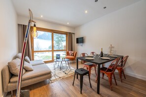 A modern living room with a sofa, dining table, and chairs, a ski rack, and a large window with a view of trees.