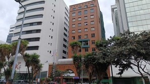 Uma rua urbana moderna com edifícios altos, incluindo um edifício branco de destaque com logotipo visível e um edifício de tijolos vermelhos com várias janelas.
