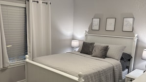 Ein Schlafzimmer mit Bett, Nachttischen, einem Fenster mit Vorhängen und Bilderrahmen an der Wand.