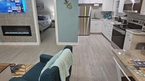 Ein modernes Wohnzimmer mit einer Couch, einem an der Wand befestigten Fernseher und einer Küchenzeile mit weißen Schränken und einem Edelstahlkühlschrank.