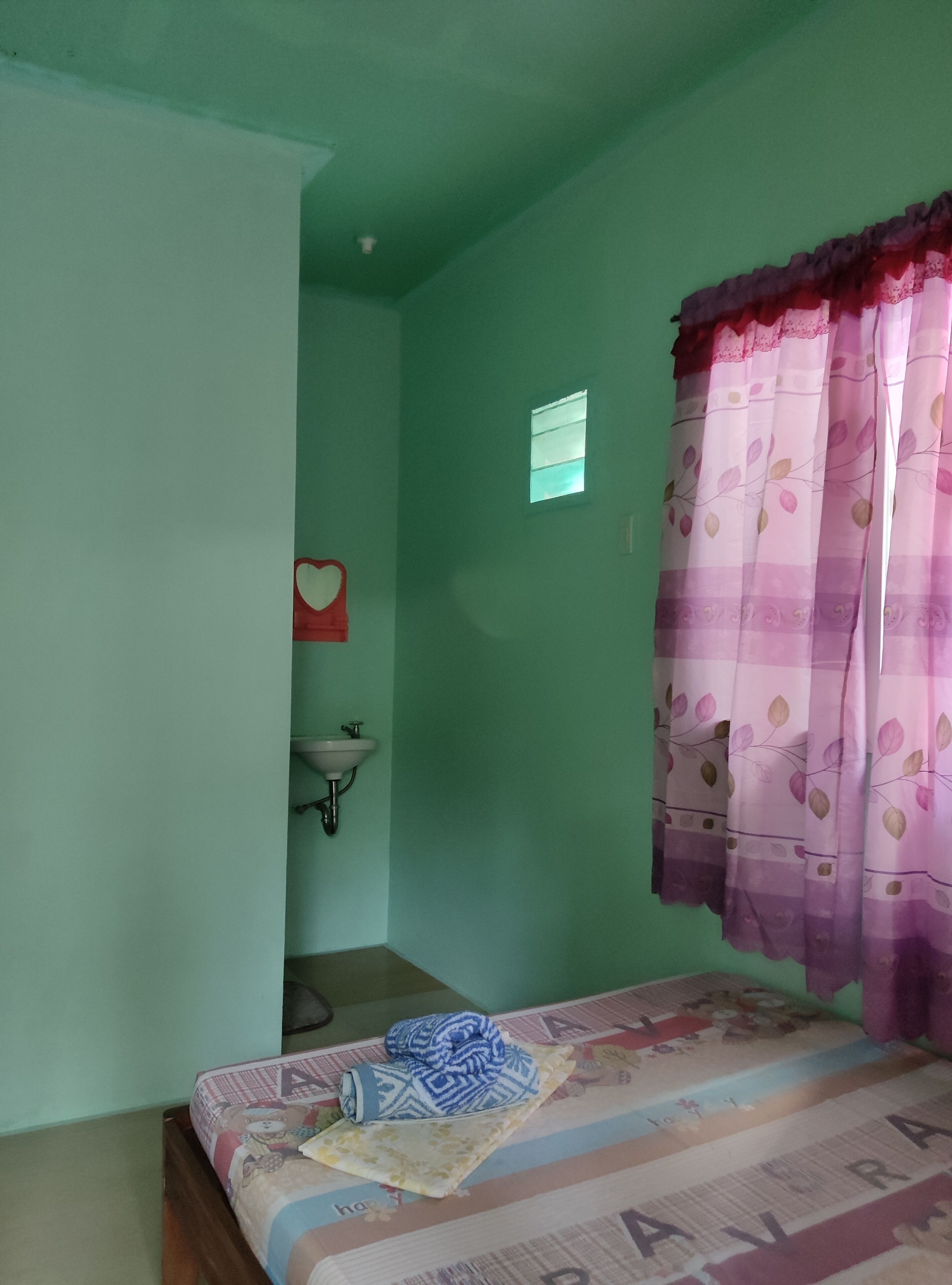 A small, single-room accommodation with a bed, a window with curtains, a sink, and a small window above the sink.