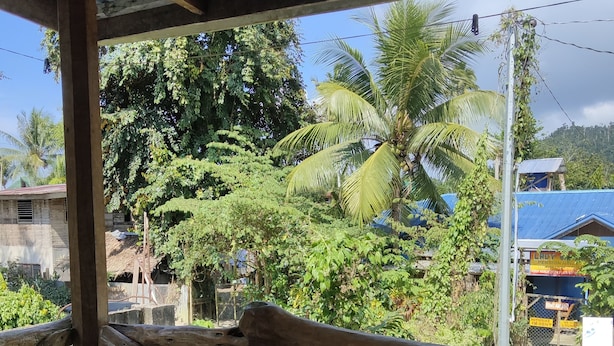 A wooden deck with a table and chairs, overlooking a lush garden with palm trees and a blue-roofed building. - Shenamae Pension  (San Vicente)