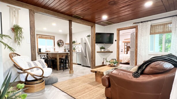 Una sala de estar acogedora con un sofá de cuero marrón, una mesa de centro de madera y una silla de mimbre. Incluye una zona de cocina con mesa y sillas, un televisor montado en la pared y un ventanal con cortinas.
