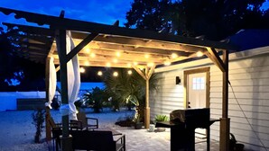 Un patio al aire libre con una pérgola, luces colgantes, una parrilla y asientos.