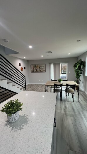 A modern kitchen with a central island, a dining area with a wooden table and chairs, and a staircase with black railings. - Normandie Hostel (Los Angeles)