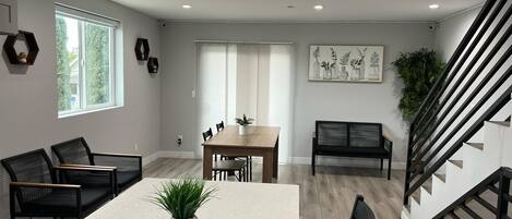 A modern living room with a dining table, chairs, a bench, a staircase, and a large window.