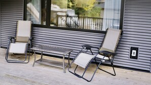 Un espace extérieur aménagé, abrité et doté de deux fauteuils et d’une petite table, offrant une vue sur un quartier résidentiel avec des arbres et une clôture.