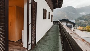 Un balcón estrecho con barandilla de madera, un edificio blanco con contraventanas oscuras y vista a un paisaje montañoso.