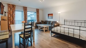 Et moderne, lyst rum med trægulv, et spisebord med stole, en sofa, en seng med metalramme og et vindue med gardiner.