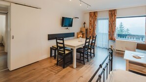 Et moderne spiseområde med et hvidt bord og sorte stole, et fladskærms-tv på væggen og et vindue med udsigt til træer.
