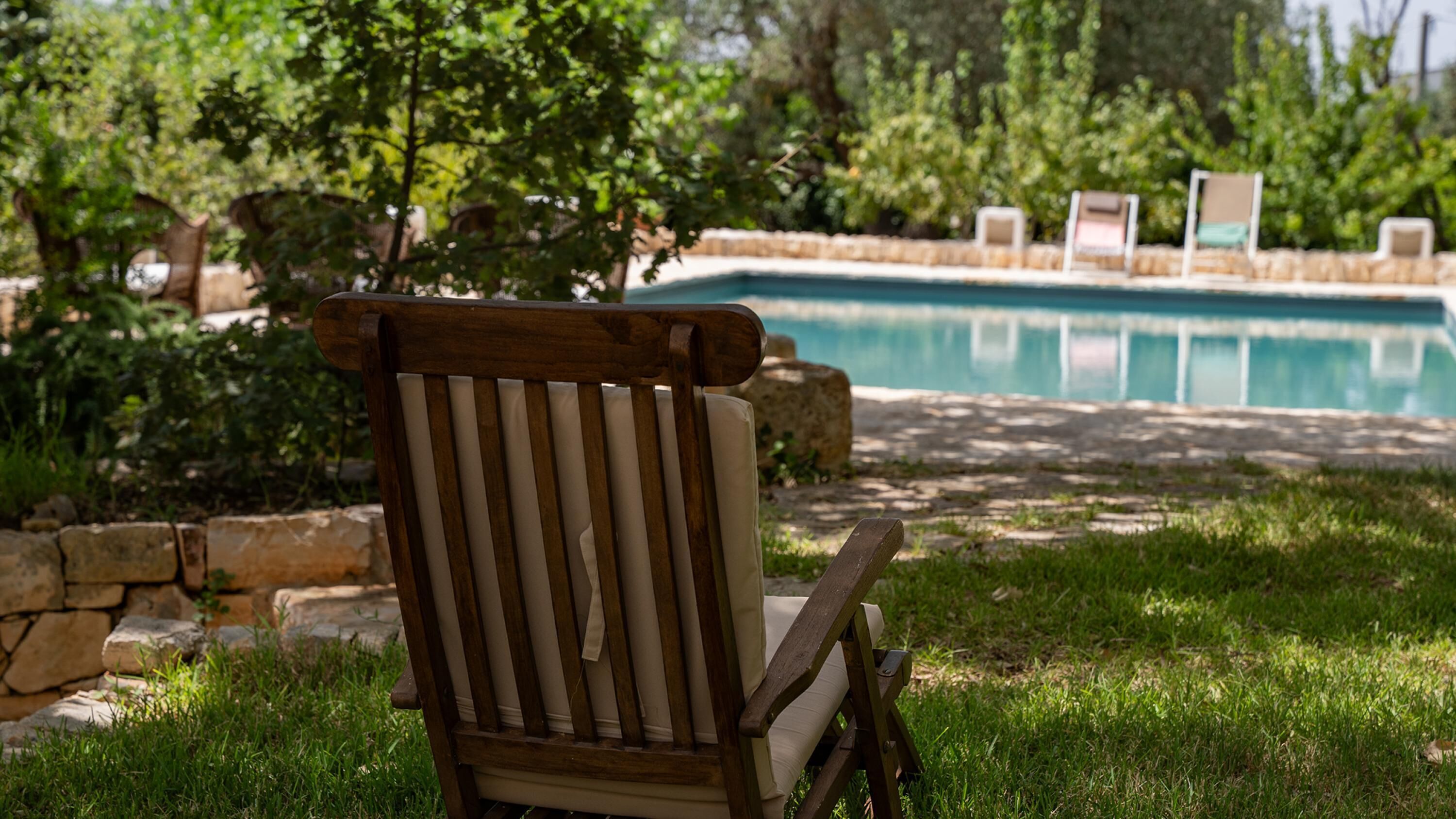 Una silla de madera con listones verticales y un cojín, ubicada frente a una piscina de agua azul cristalina, rodeada de vegetación y muros de piedra.