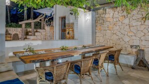 A dining area with a long wooden table, wicker chairs, a stone wall, and a hanging vine. - Villa Giari 8 Emma Villas (Ceglie Messapica)