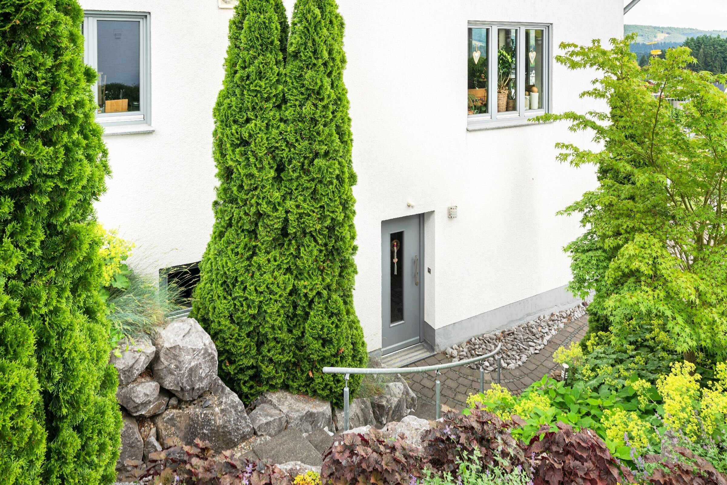 A white building with a door and windows, surrounded by greenery and a stone pathway.