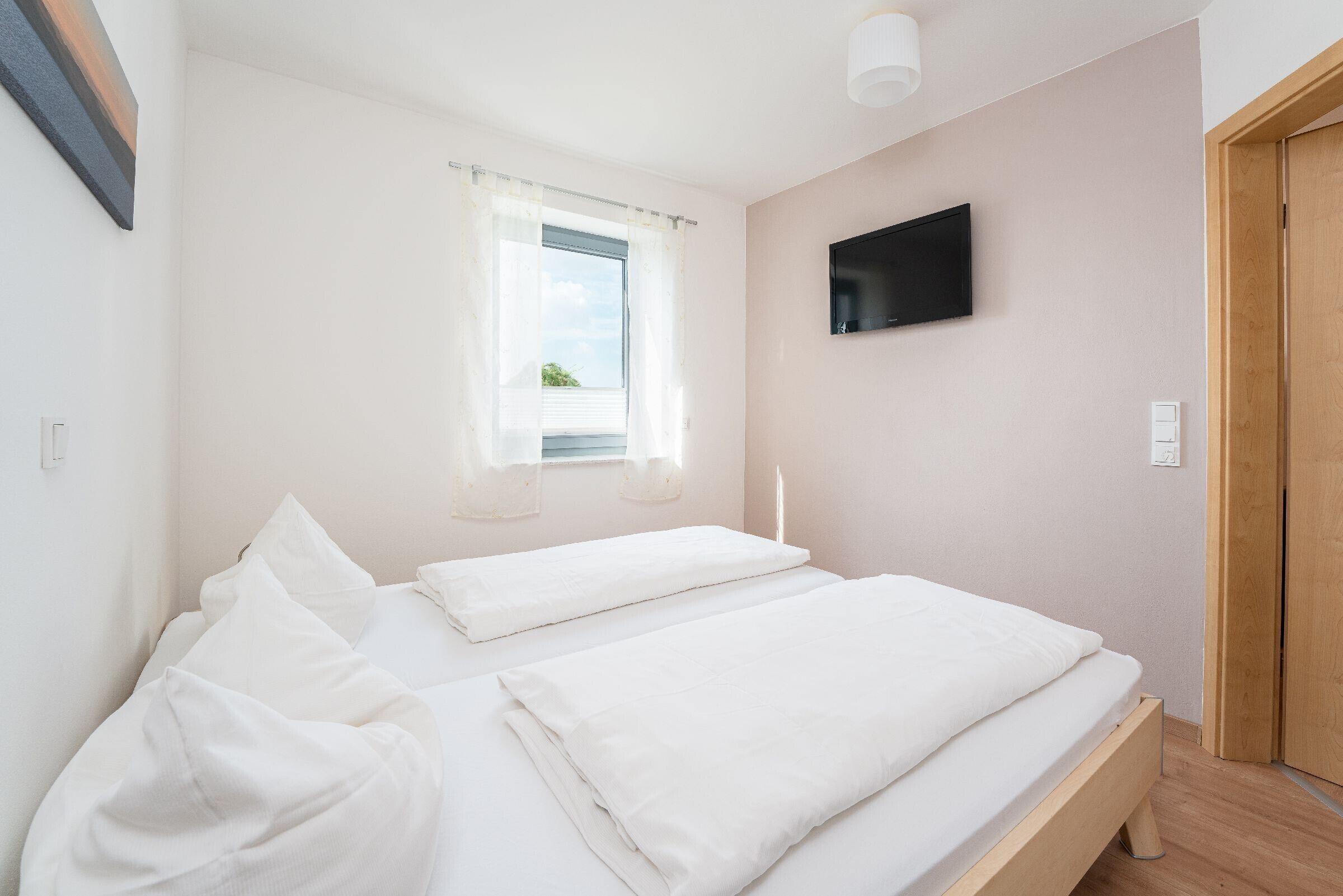 Ein Schlafzimmer mit zwei Betten, einem Fenster mit Vorhängen, einem Flachbildfernseher an der Wand und einer Holztür.