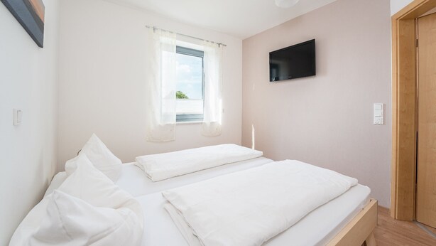 Ein Schlafzimmer mit zwei Betten, einem Fenster mit Vorhängen, einem Flachbildfernseher an der Wand und einer Holztür.