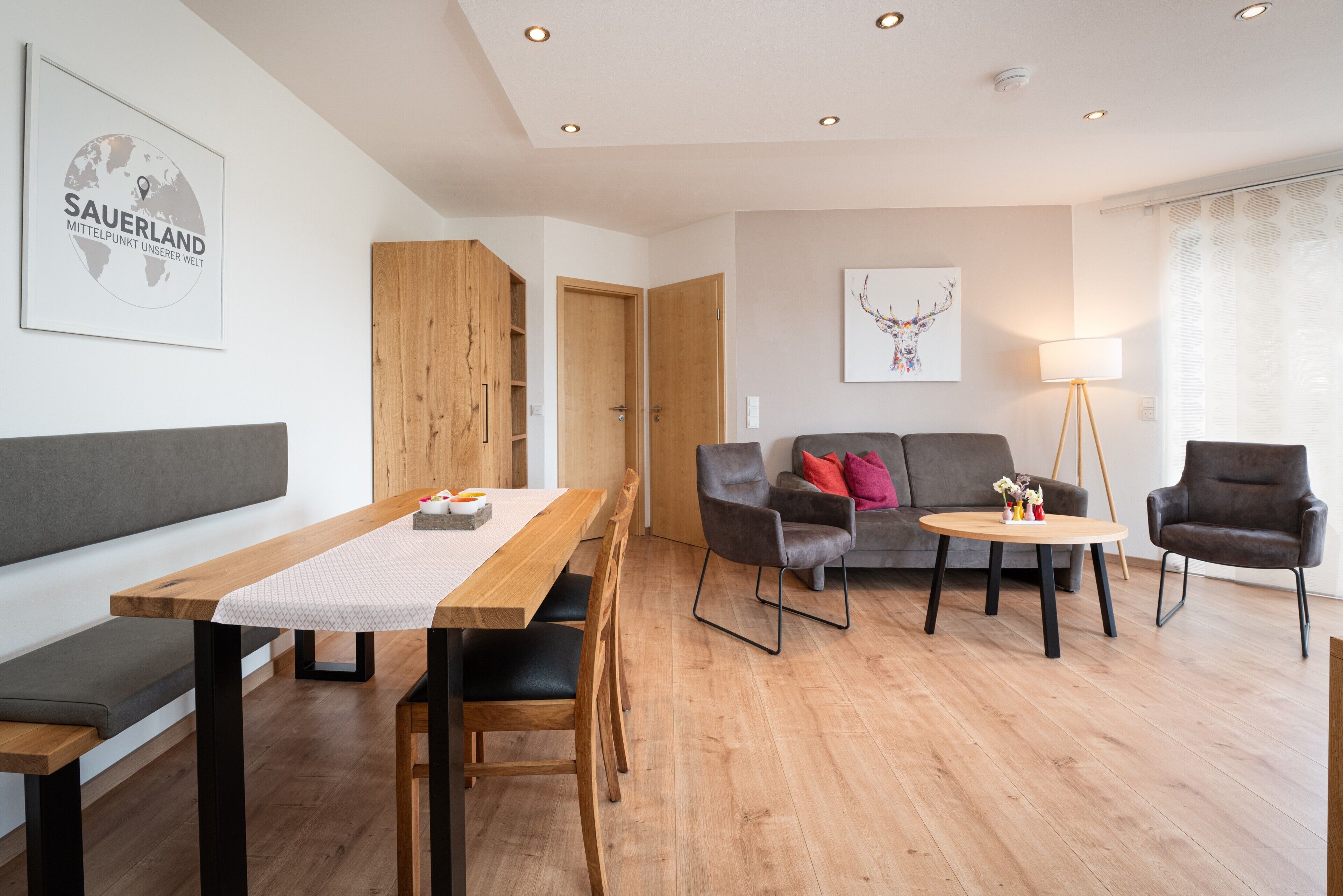 A modern living room with a wooden dining table, grey sofa, and a small round table with a lamp.