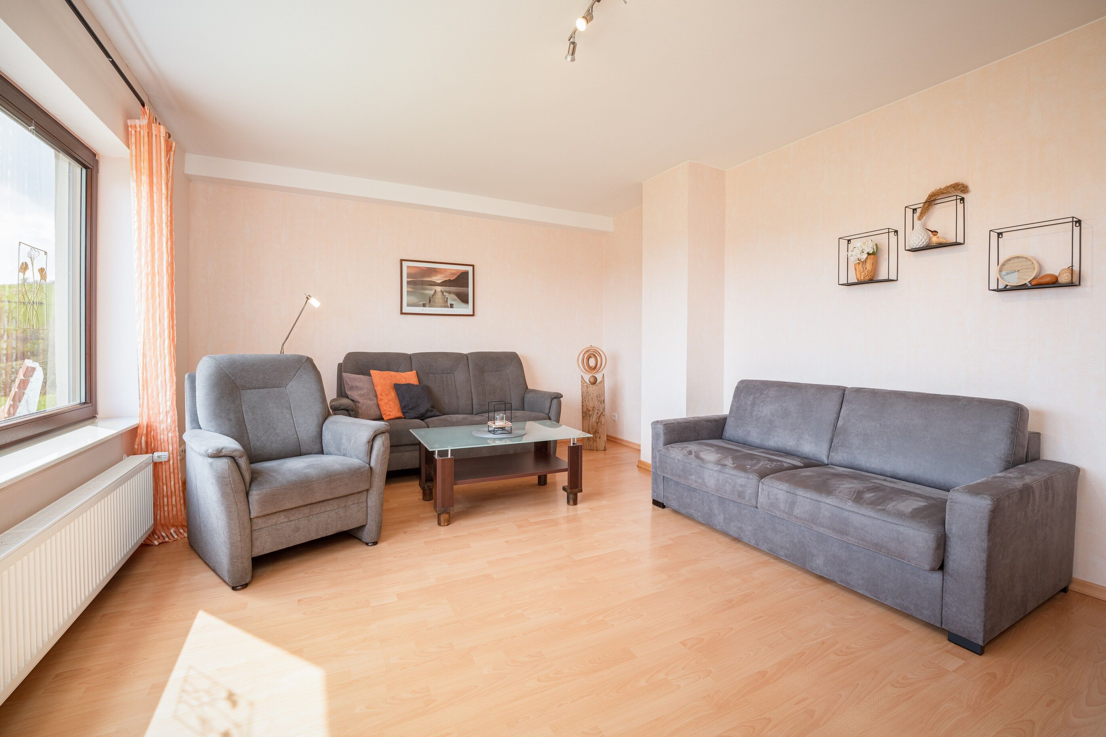 Un salón con sofá gris, un sillón gris, una mesa de centro y una lámpara de pie. Se ve una ventana con cortinas, un radiador y un cuadro en la pared.