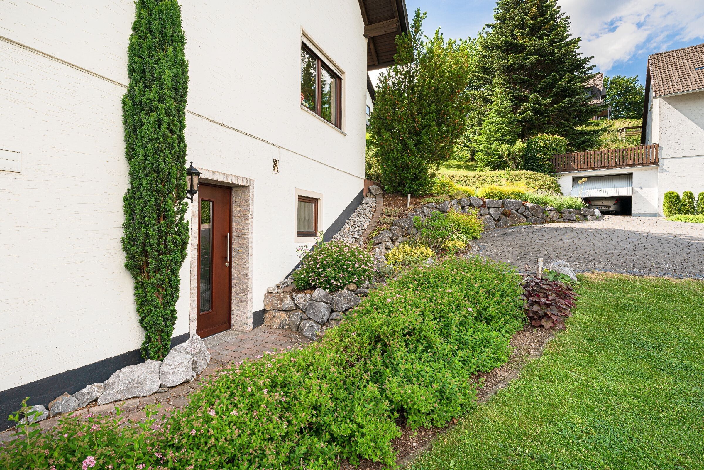 A white building with a stone staircase, a garage, and a well-maintained garden with various plants and flowers.