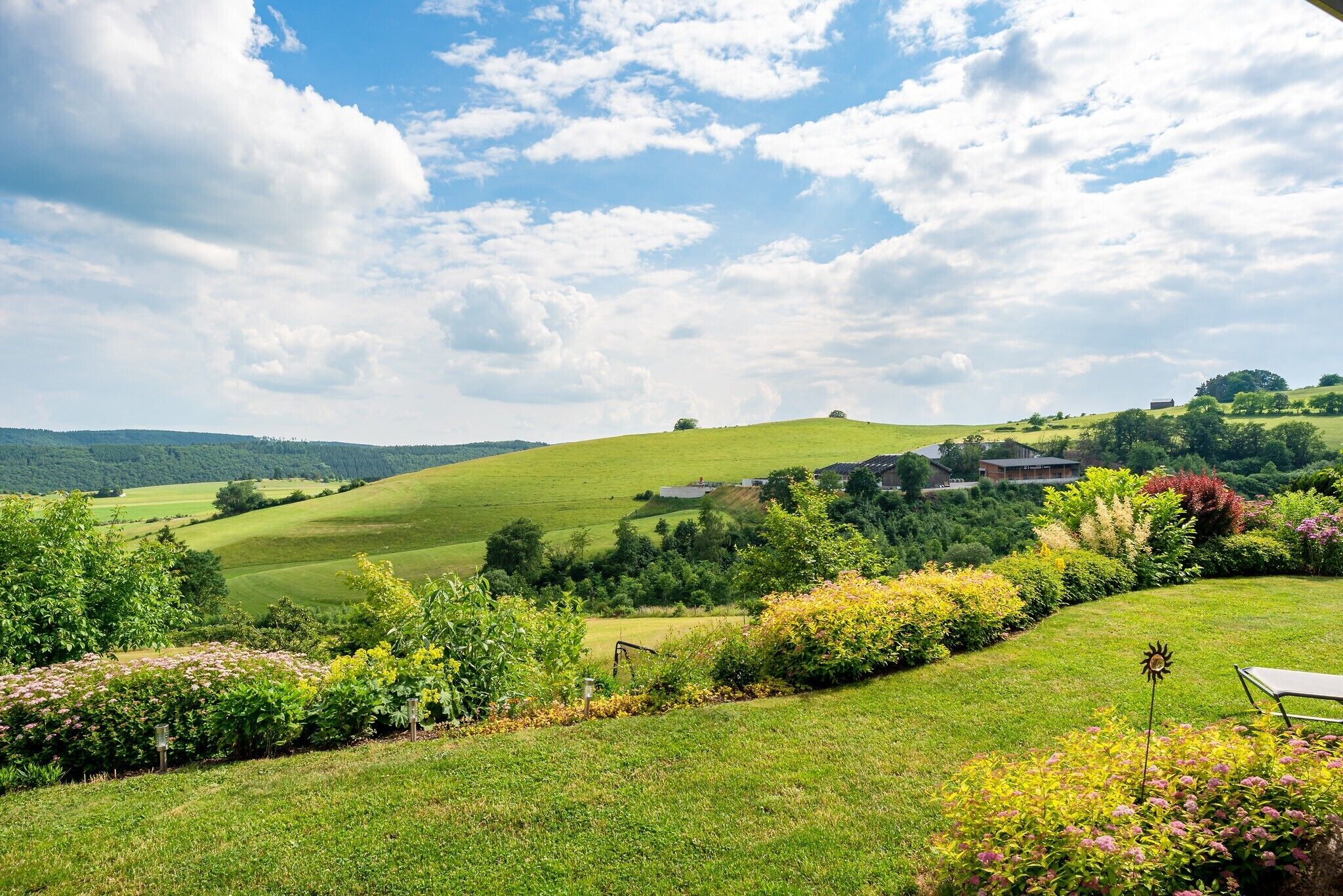 Ländliche Landschaft mit sanften Hügeln, einem kleinen Haus und vielfältiger Vegetation.