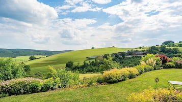 Ländliche Landschaft mit sanften Hügeln, einem kleinen Haus und vielfältiger Vegetation.