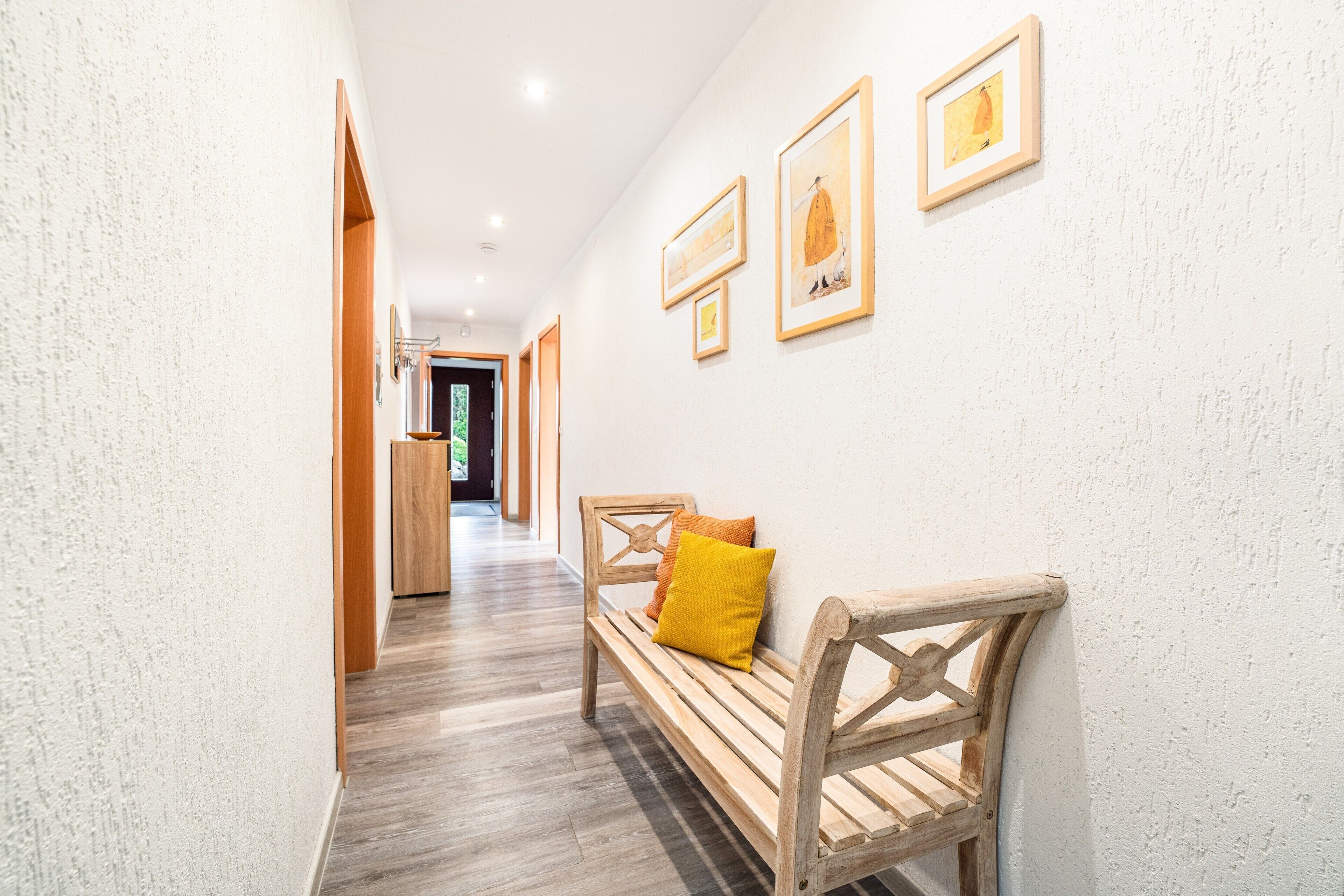 A hallway with a wooden bench, framed artwork on the wall, and a wooden door at the end.