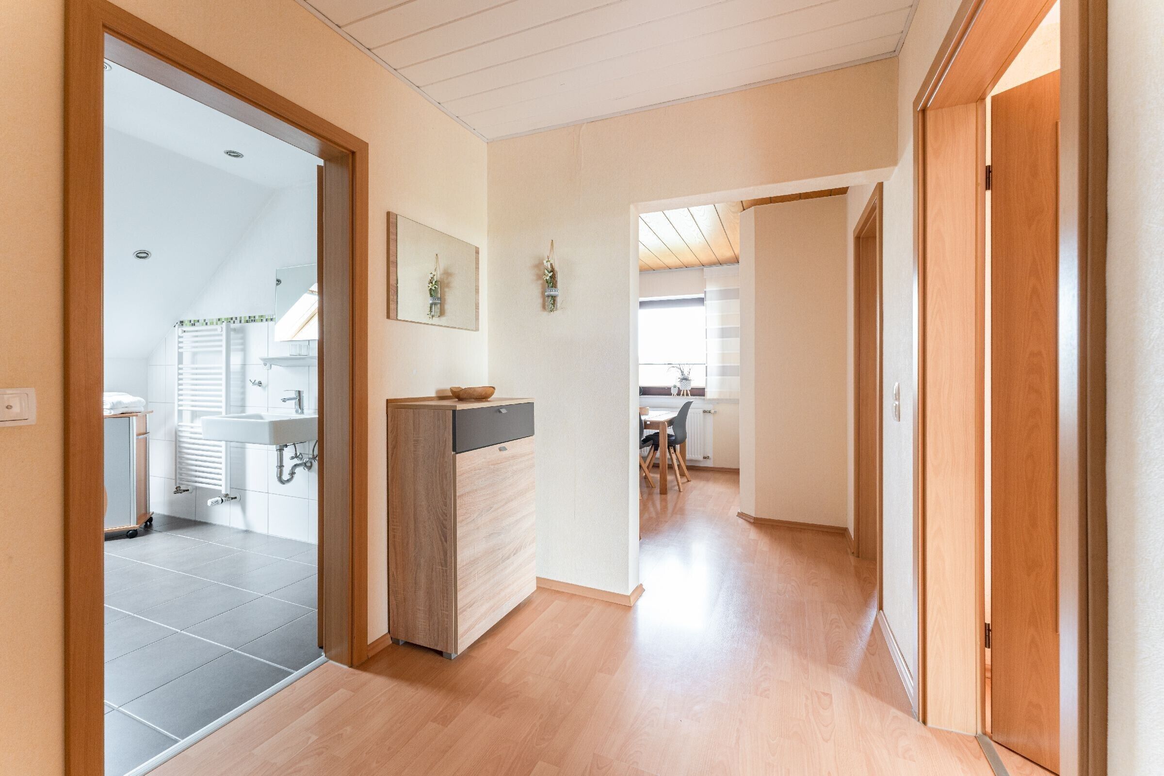 A hallway with a wooden floor, a kitchen area with a sink and cabinets, and a dining area with chairs and a table.