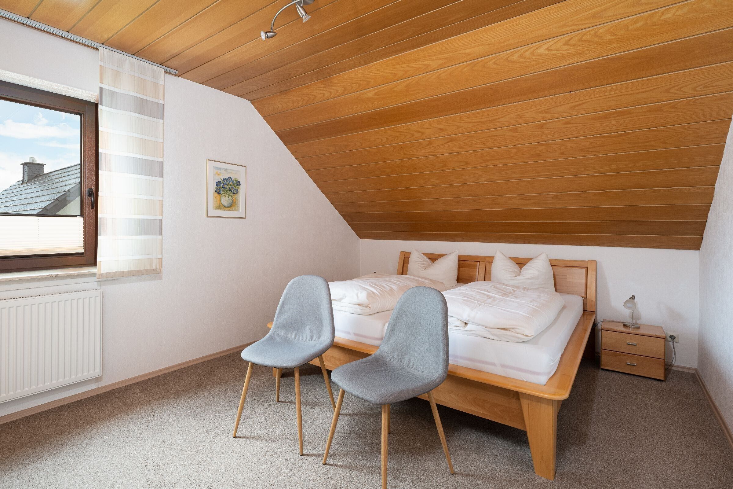 A bedroom with a sloped ceiling, a bed, two chairs, a nightstand, and a window.