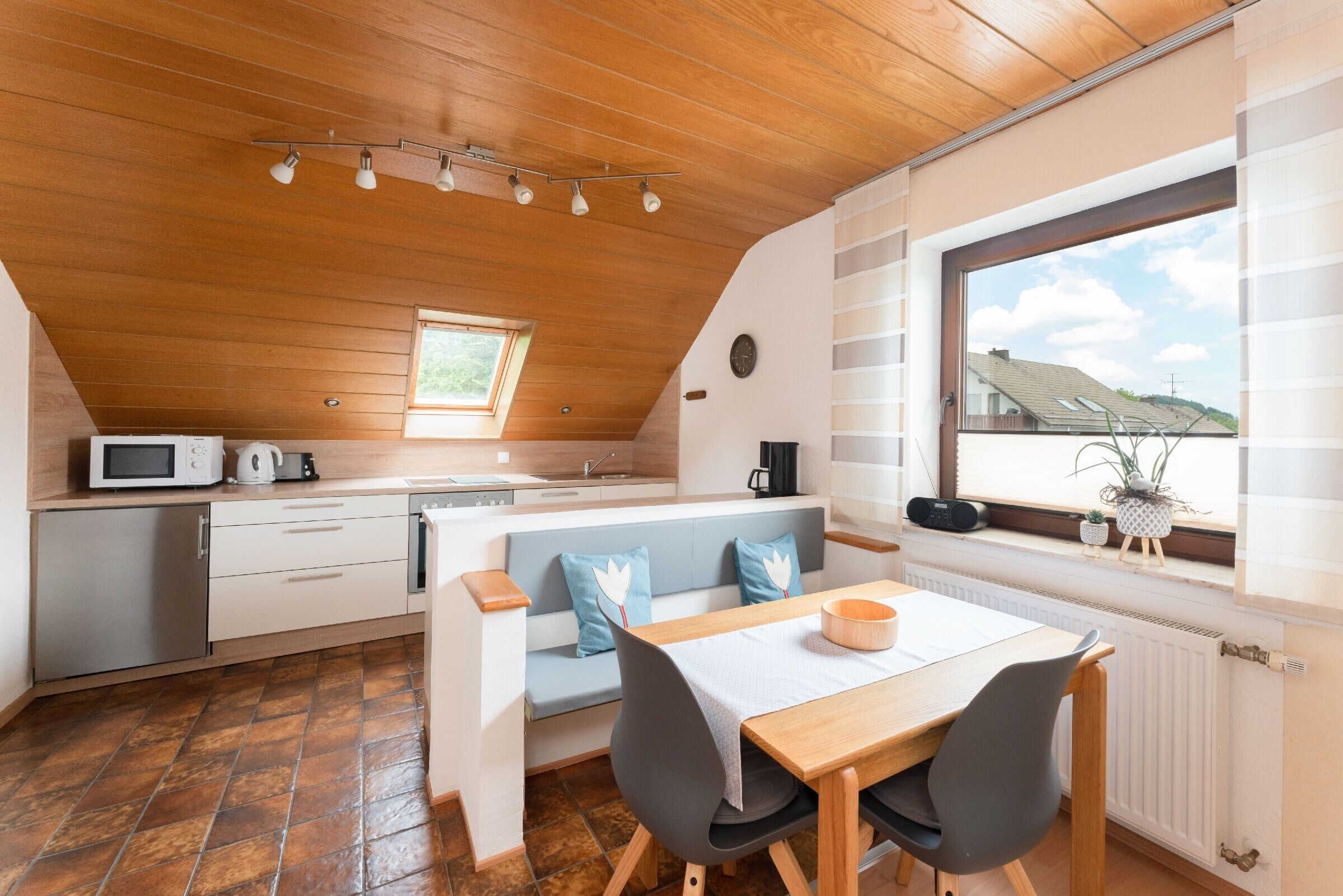 A modern kitchen with a wooden ceiling, a dining area with a table and chairs, a built-in bench, and a window with a view of houses.