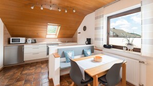 A modern kitchen with a wooden ceiling, a dining area with a table and chairs, a built-in bench, and a window with a view of houses.