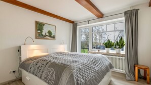 Une chambre à coucher avec un lit, une lampe de chevet, une fenêtre avec des rideaux, un radiateur et un petit tabouret en bois.