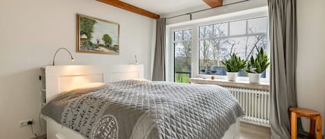 A bedroom with a bed, bedside lamp, window with curtains, radiator, and a small wooden stool.