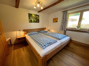 Ein Zimmer mit zwei Betten, Holzboden, einer Deckenleuchte, einem Fenster mit Vorhängen und einem Heizkörper.
