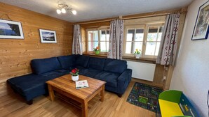 Une pièce chaleureuse avec un plancher en bois, un canapé bleu, une table basse en bois sur laquelle reposent un livre et des fleurs, une fenêtre avec des rideaux et un radiateur.