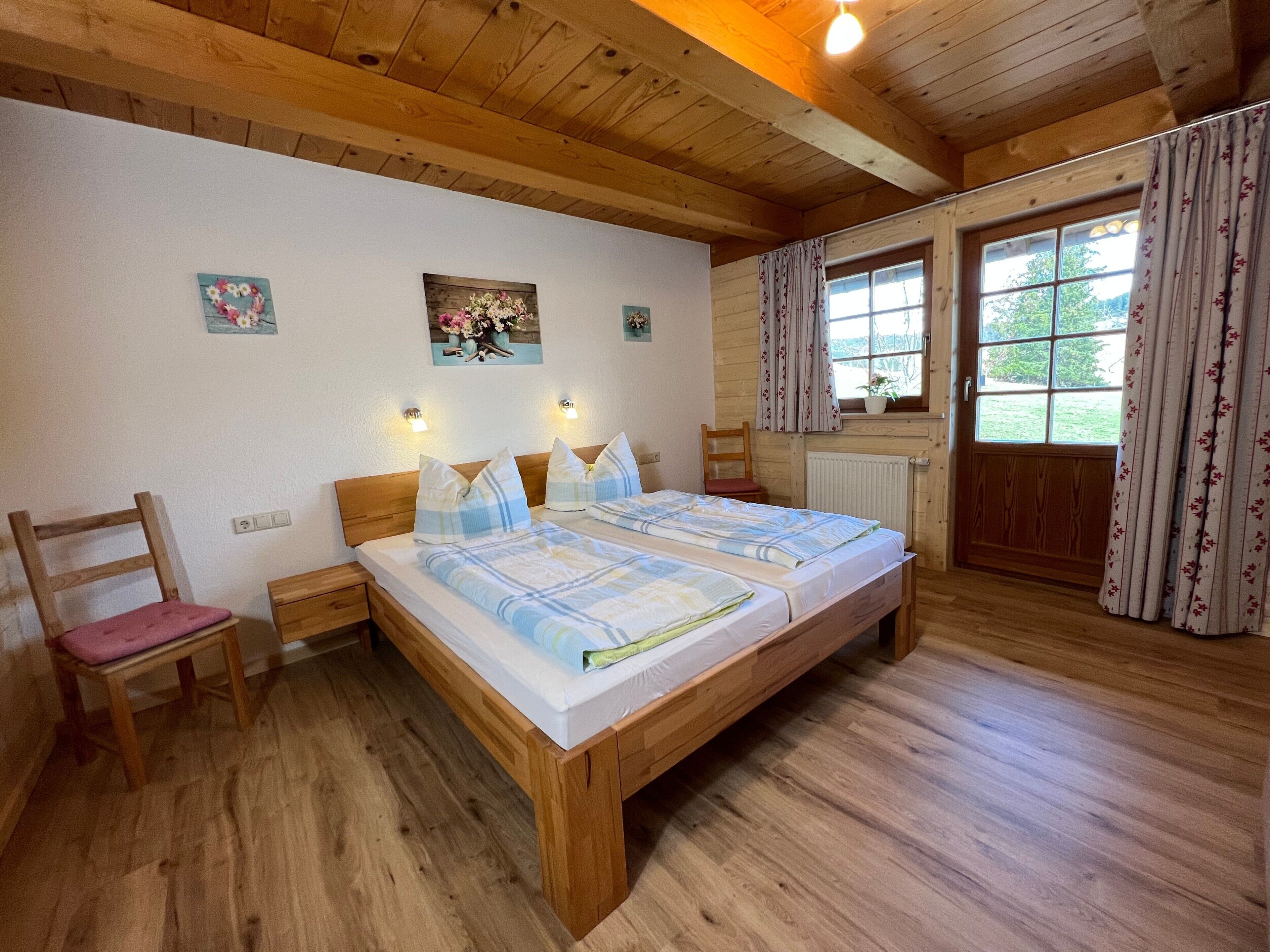 A wooden-framed bedroom with a bed, two bedside tables, a chair, a window with curtains, and a wall-mounted light fixture.