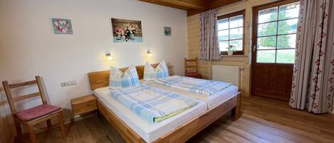 A wooden-framed bedroom with a bed, two bedside tables, a chair, a window with curtains, and a wall-mounted light fixture.