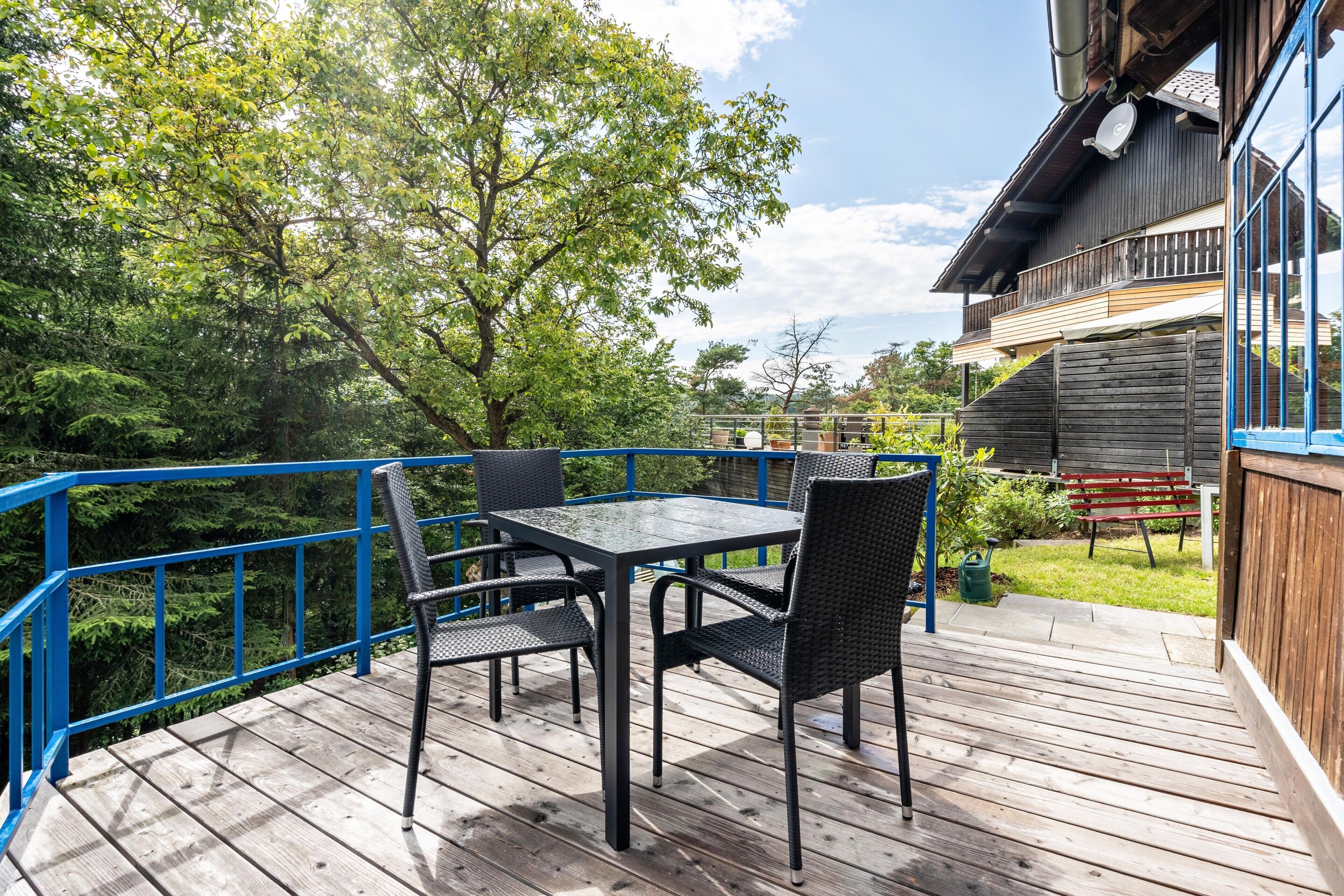 Eine Holzterrasse mit Tisch und Stühlen, umgeben von einem blauen Geländer, mit Blick auf einen Garten und ein Gebäude.
