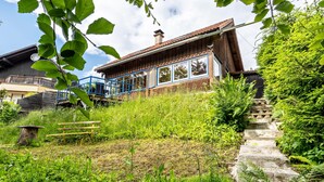 Une maison en bois à l’allure rustique, dotée d’un balcon et entourée d’une végétation luxuriante, avec un escalier en pierre.