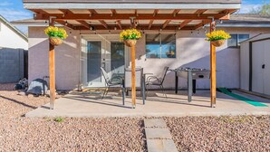 Un patio couvert avec une table et des chaises, trois jardinières suspendues et une structure en forme de pergola.