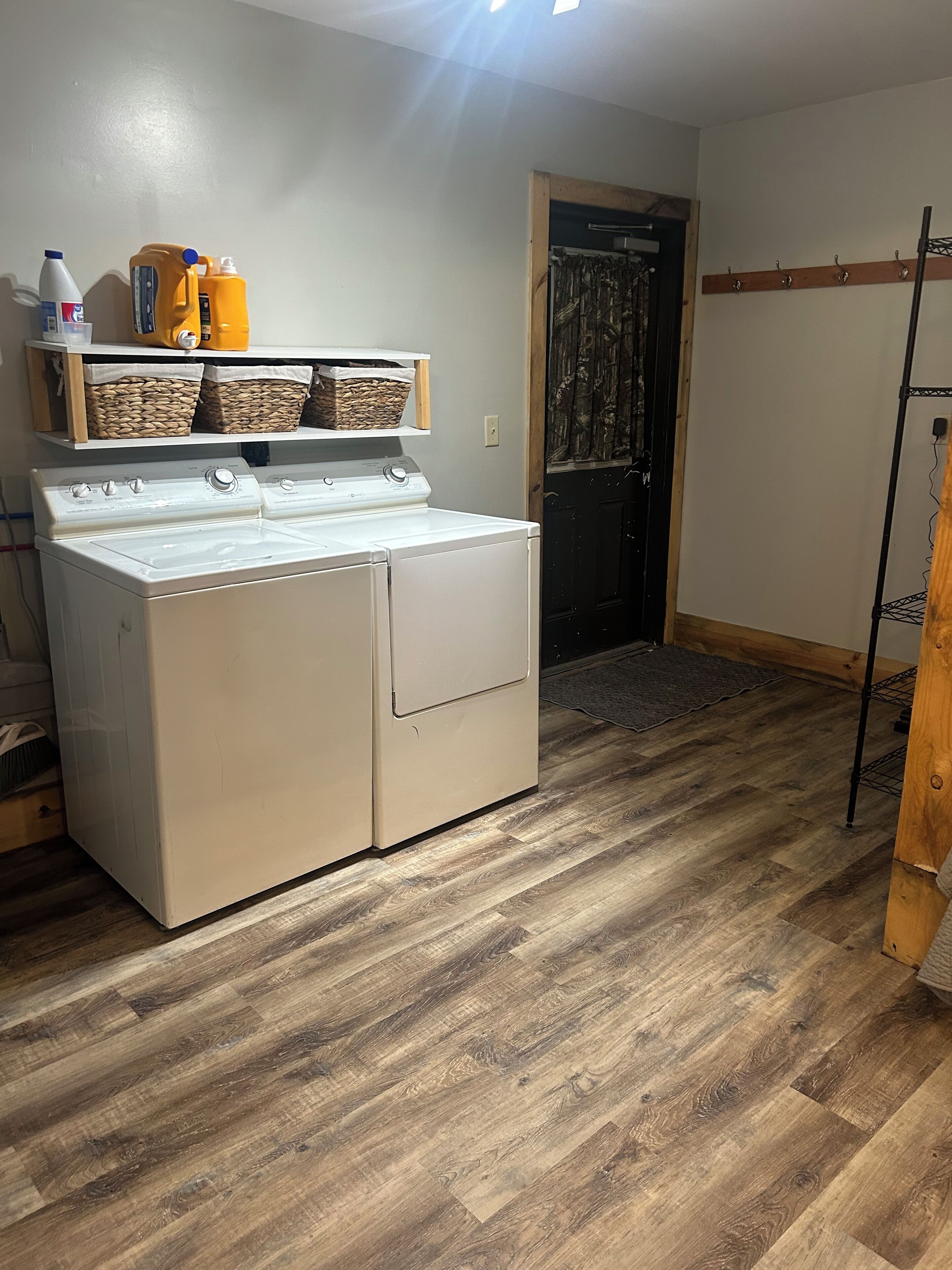 Une buanderie équipée d’un lave-linge et d’un sèche-linge à chargement frontal, d’un plancher en bois, d’une porte avec un rideau et d’étagères contenant des paniers et des produits de nettoyage.