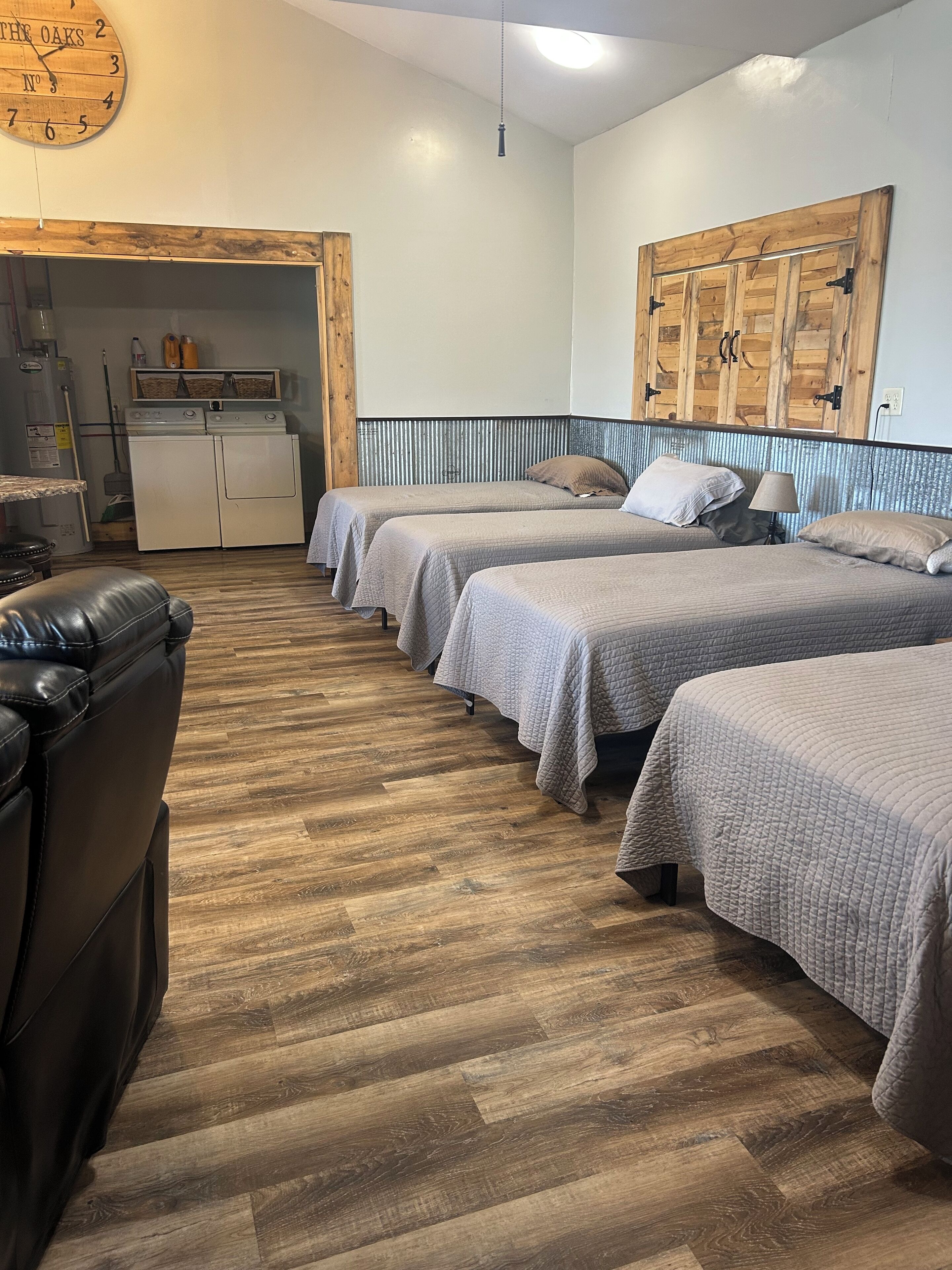 A hotel room with three beds, a wooden floor, a clock on the wall, and a kitchenette area.