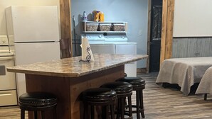 Una cocina con isla de madera y taburetes, un refrigerador, una estufa y una cama al fondo.