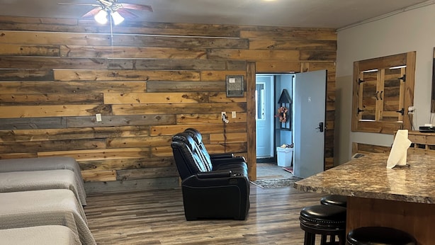 Una habitación con piso de madera, ventilador de techo, una butaca de cuero negro, una zona de bar con taburetes y una puerta que da a otra habitación.