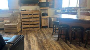 A spacious living area with a wooden floor, a leather sofa, a wooden coffee table, and a kitchen area with a microwave and bar stools.