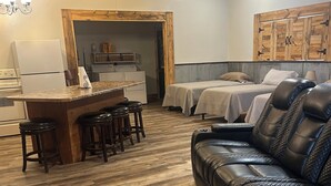 Una sala de estar acogedora con piso de madera, un sofá de cuero negro, una barra de madera con taburetes y una cama con dos almohadas.