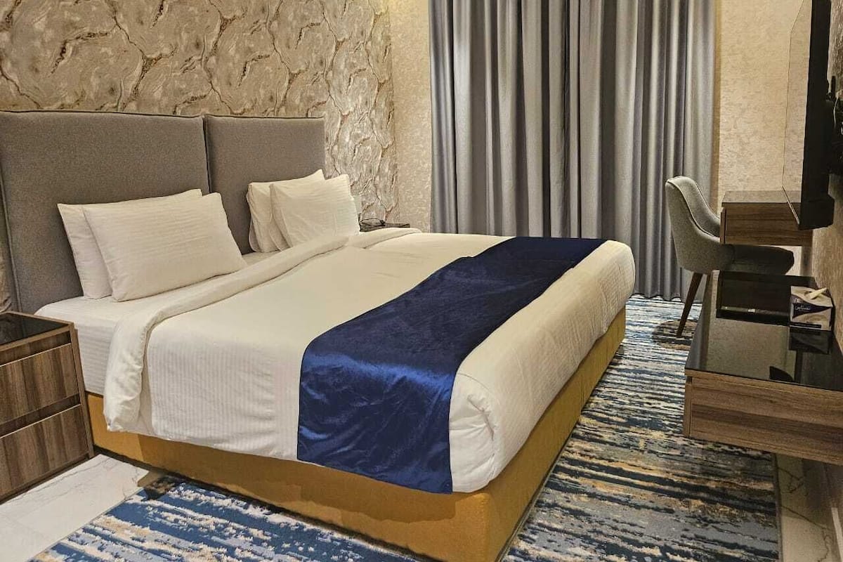 Una habitación de hotel con una cama grande, una mesita de noche de madera, un escritorio con una silla y una pared con textura de piedra.